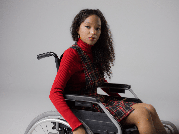 A disabled teenage girl, wearing lipstick to match her autumnal high neck top and tartan dress, looks at the camera with a neutral expression from her wheelchair in a studio