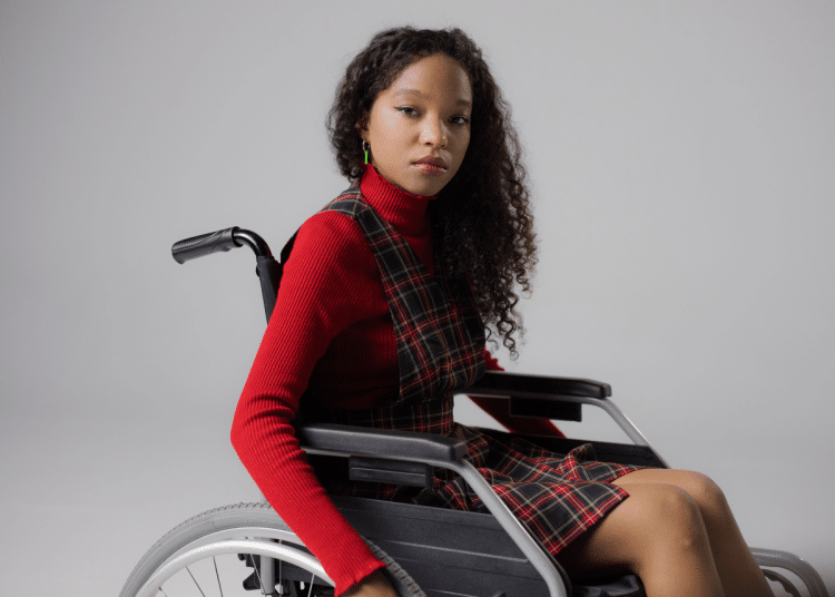 A disabled teenage girl, wearing lipstick to match her autumnal high neck top and tartan dress, looks at the camera with a neutral expression from her wheelchair in a studio