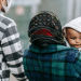 Asylum seeker story: A couple walking outside and the woman is holding a toddler. Only his face is visible.