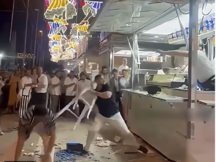 A screenshot of the video showing a pair of Israeli men, allegedly intoxicated, with one holding a plastic stool in his hand about to launch it at the stall and its three staff members