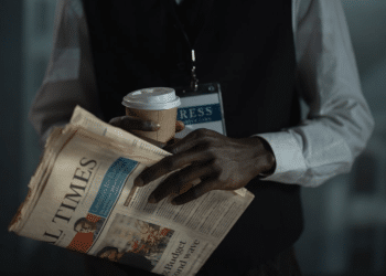 The torso of a male journalist holding a copy of the Financial Times, with a cardboard cup holding a drink, and you can see a press card attached to a lanyard around his neck. His neck upwards is not visible