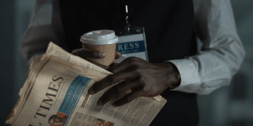 The torso of a male journalist holding a copy of the Financial Times, with a cardboard cup holding a drink, and you can see a press card attached to a lanyard around his neck. His neck upwards is not visible