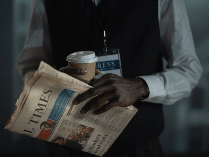The torso of a male journalist holding a copy of the Financial Times, with a cardboard cup holding a drink, and you can see a press card attached to a lanyard around his neck. His neck upwards is not visible