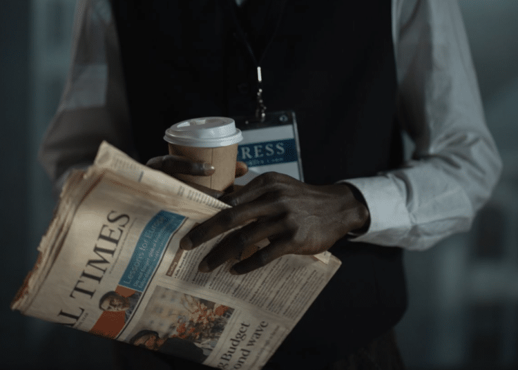 The torso of a male journalist holding a copy of the Financial Times, with a cardboard cup holding a drink, and you can see a press card attached to a lanyard around his neck. His neck upwards is not visible