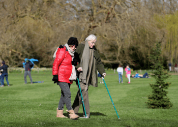 Carers Week: An older pair, one with two crutches, and the other locking their arm, walk and enjoy the spring sunshine at Mottisfont one of the National Trust parklands on 21 March 2020 in Romsey, UK