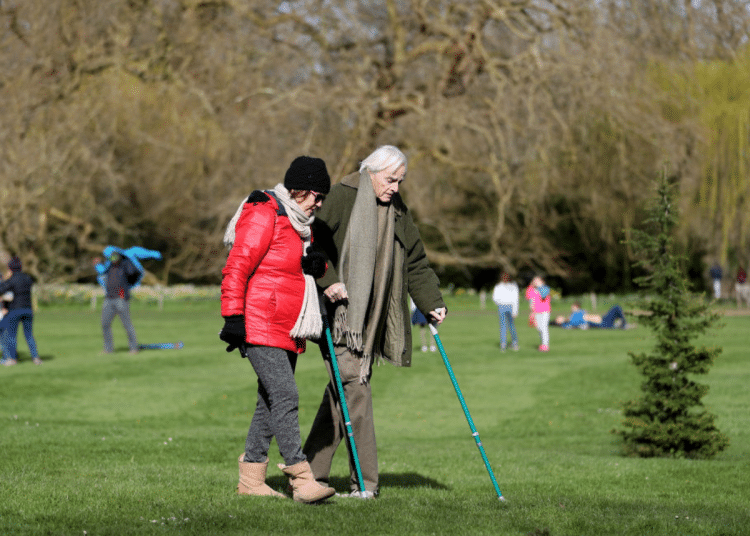 Carers Week: An older pair, one with two crutches, and the other locking their arm, walk and enjoy the spring sunshine at Mottisfont one of the National Trust parklands on 21 March 2020 in Romsey, UK