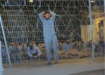 A leaked photograph of the Sde Teiman detention facility shows a blindfolded Palestinian man with his arms above his head behind a metal wire fence in 2024. Knesset politicians defeated a bill to ban visits from the Red Cross.