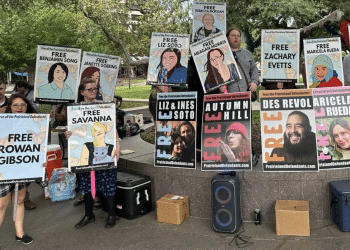Posters with 'Free XYZ' line a wall and one woman holds one saying 'Free Rowan Gibson'. They are part of the protestors jailed for demonstrating outside an ICE centre