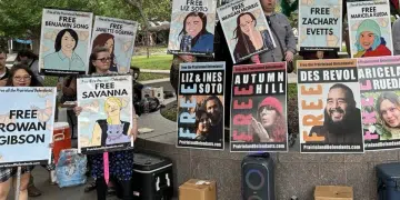 Posters with 'Free XYZ' line a wall and one woman holds one saying 'Free Rowan Gibson'. They are part of the protestors jailed for demonstrating outside an ICE centre