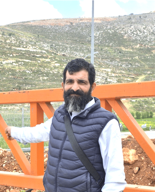 Ayed Ghafari, a human rights activist, who lives in Sinjil, Palestine, smiles infront of a large gate that is outside. Behind him are high grassy hills