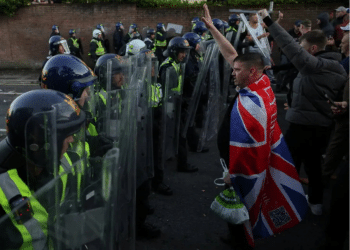 Armed riot police hold their shields up in a row while men, one at the front with Union Jack flag draped on his back, show Nazi salutes. Skwawkbox argues Israel-bought politicians show hypocrisy when responding to riots and protests