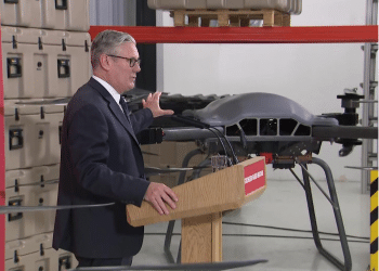 Keir Starmer touching the top of a drone inside a company factory, speaking to a crowd at a wooden podium. We cannot see the crowd.