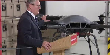 Keir Starmer touching the top of a drone inside a company factory, speaking to a crowd at a wooden podium. We cannot see the crowd.