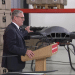 Keir Starmer touching the top of a drone inside a company factory, speaking to a crowd at a wooden podium. We cannot see the crowd.