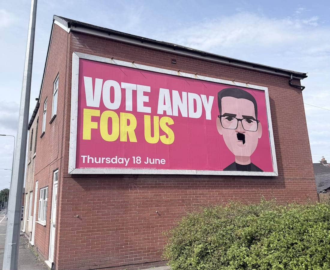 A billboard saying 'Vote Andy For Us' with a cartoon image of Andy Burnham, which has been altered to give him a Hitler-style moustache.