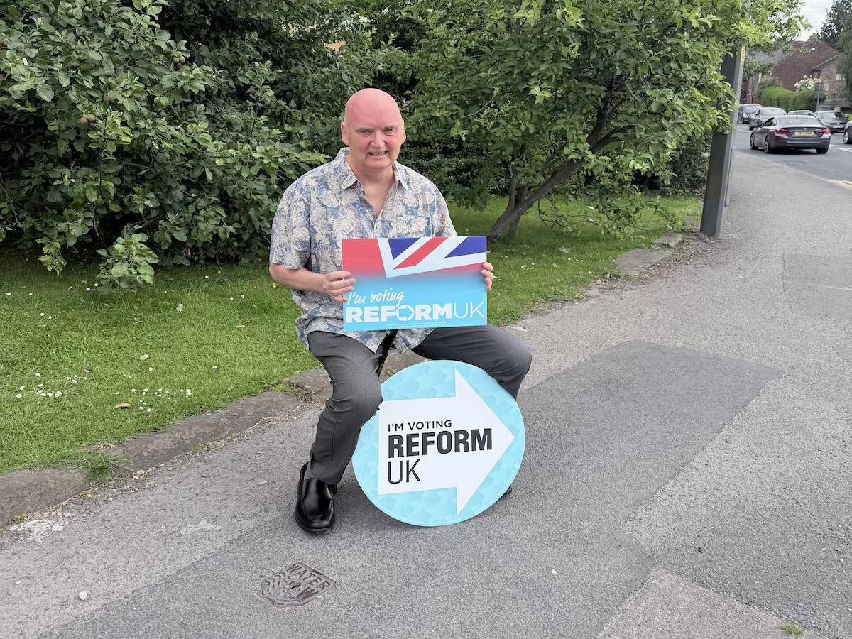 An older man crouches down and almost squats above a Reform placard