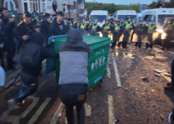 A still from a video posted on Young Bob's X account of two males pushing a large wheelie bin with a fire inside towards a row of armed riot police at far-right riots this week. Others line the pavements filming with their mobile phones and cheering the pair on