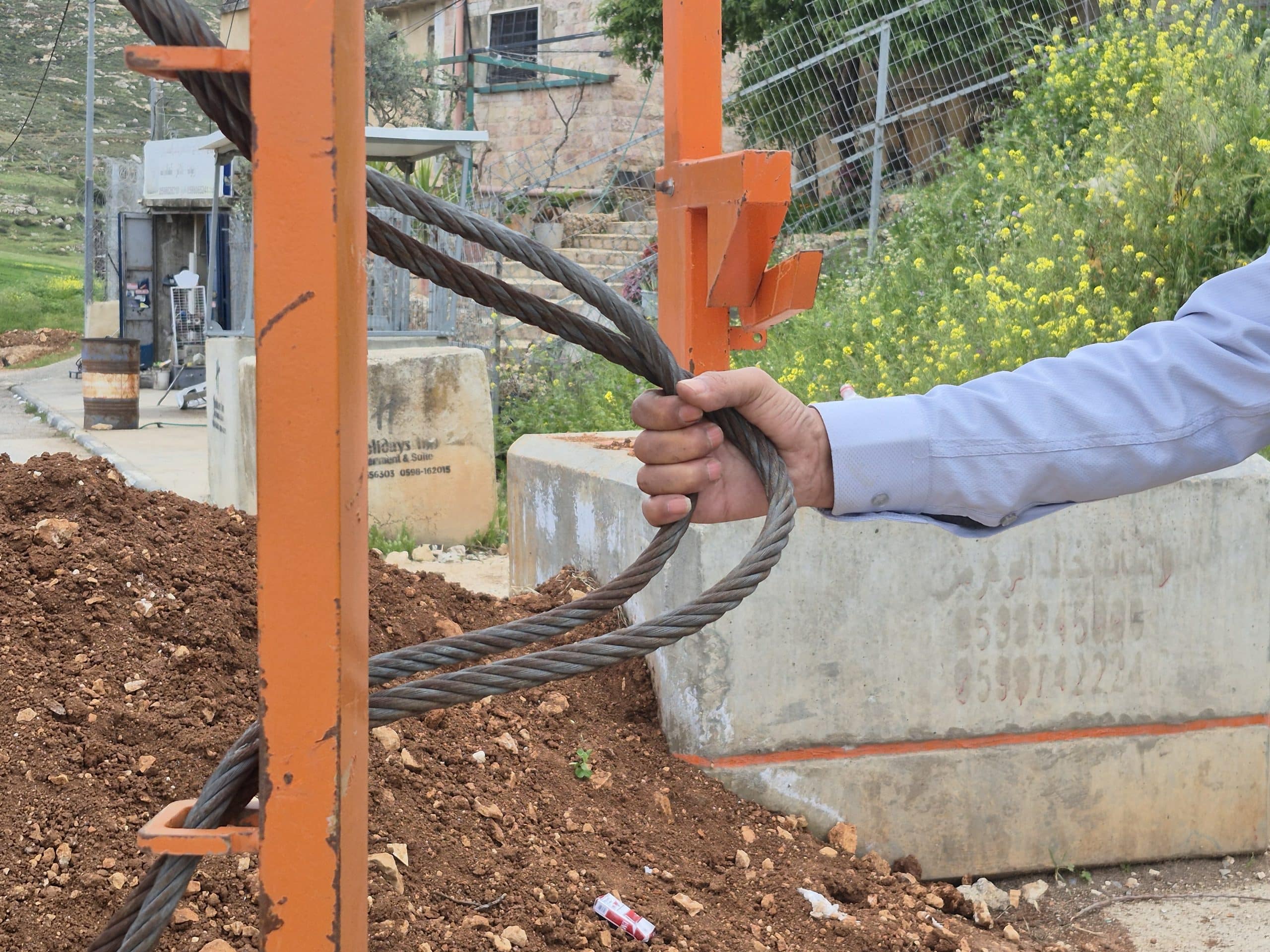 Ayed Ghafari's hand grips a pair of thick, dirty cable wires threaded around the gate post. It's a close-up of his hand and arm only