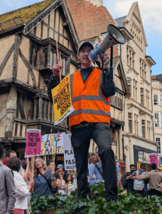 A man with a megaphone on top of a planter. He is wearing a bright orange hi-vis and is holding a Stand Up To Racism sign upside down