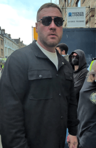 Large heavy set white man in a black jacket and dark aviators looks at the camera threateningly Tommy Robinson