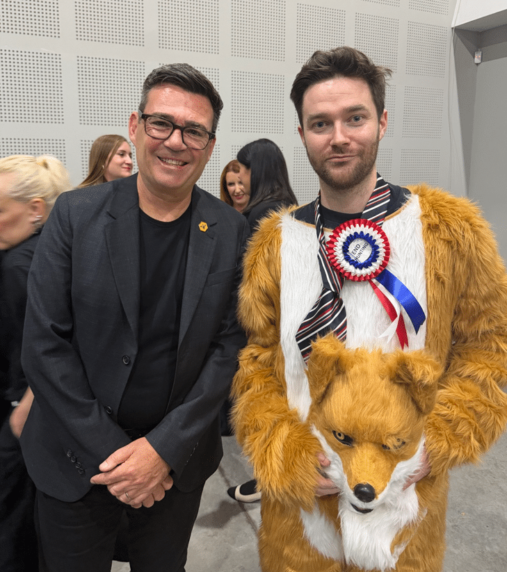 Andy Burnham (left) leans into Rob Pownall at the Makerfield by-election count, where they pose for a photo. Burnham is wearing a suit blazer with crew neck tshirt, smiling. Pownall is dressed as a red fox, but he has taken the mask off. His face looks amused.
