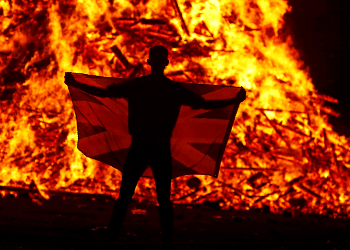 A male holds a flag in front of the Loyalist Corcrain Redmanville bonfire, which was lit to mark the start of the unionist 12 July celebrations, in Portadown, Northern Ireland, on 11 July 2021.