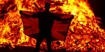 A male holds a flag in front of the Loyalist Corcrain Redmanville bonfire, which was lit to mark the start of the unionist 12 July celebrations, in Portadown, Northern Ireland, on 11 July 2021.