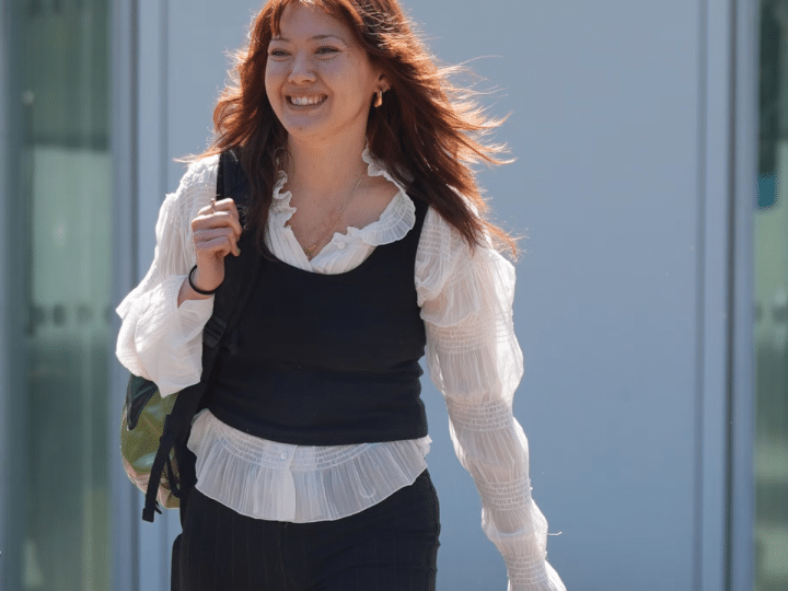 Ellie Kamio, one of the Filton 25, smiling while walking outside Woolwich Crown Court, London, in April 2026 with one hand on her shoulder keeping her backpack strap in place