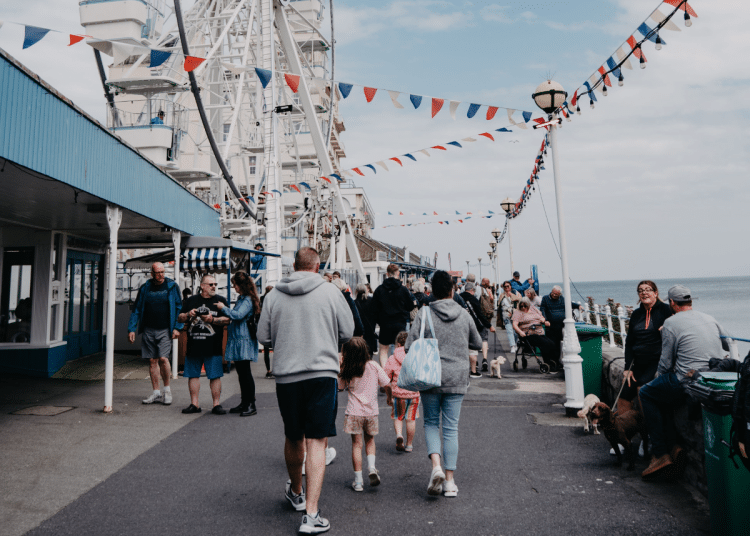 A bustling scene at Llandudno promenade in Wales with people walking near a Ferris wheel adorned with flags. Days out like this will not be affordable for many UK families due to money worries.