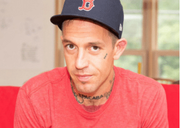 Fergie Chambers, who has small face tattoos and a neck tattoo on show above his crew neck tshirt, looks at the camera from beneath his baseball cap. He is a vocal supporter of Palestine.