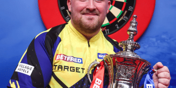 Luke "the Nuke" Littler smiles with his winner's trophy in hand infront of a darts board in Blackpool