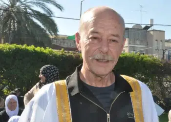 Maher Younis, fomer Palestinian prisoner, has a full grey moustache and bald head. He smiles, the sun shining on him and the white/ gold-trimmed robe over his black shirt
