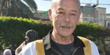 Maher Younis, fomer Palestinian prisoner, has a full grey moustache and bald head. He smiles, the sun shining on him and the white/ gold-trimmed robe over his black shirt