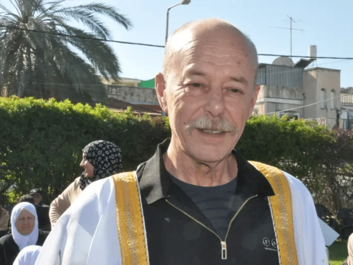 Maher Younis, fomer Palestinian prisoner, has a full grey moustache and bald head. He smiles, the sun shining on him and the white/ gold-trimmed robe over his black shirt