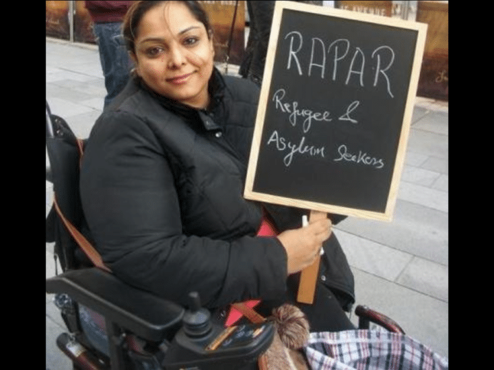 Manjeet Kaur, a human rights campaigner who died, aged 41, in 2020 in Manchester, sought asylum in the UK from Afghanistan in March 2011. In this photo in her wheelchair, she’s smiling holding a chalkboard placard that says 'RAPAR - Refugees and Asylum Seekers'.
