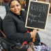 Manjeet Kaur, a human rights campaigner who died, aged 41, in 2020 in Manchester, sought asylum in the UK from Afghanistan in March 2011. In this photo in her wheelchair, she’s smiling holding a chalkboard placard that says 'RAPAR - Refugees and Asylum Seekers'.