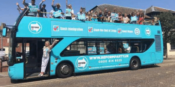 A Reform branded open top double decker bus parked with people on top waving and a woman in the doorway waving too, in 2013