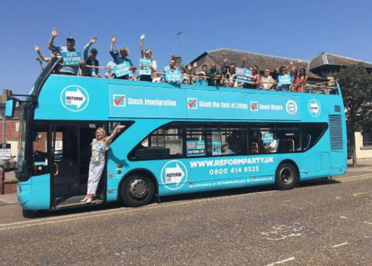 A Reform branded open top double decker bus parked with people on top waving and a woman in the doorway waving too, in 2013