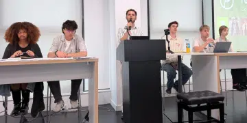 Young Greens party conference. Each candidate is sat at the front of a classroom on two desks with a podium and mic in the middle, where one candidate is speaking